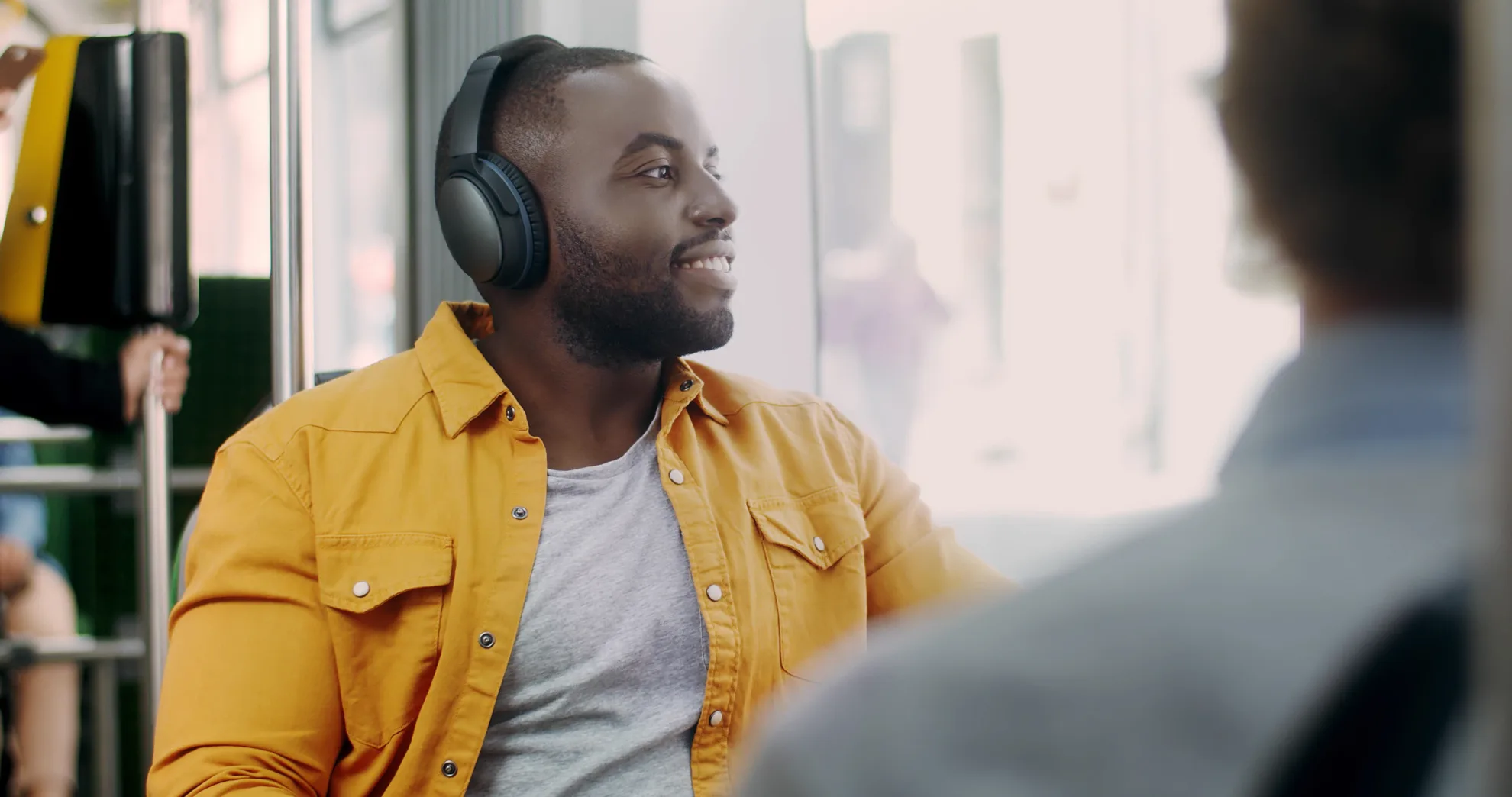 Smiling young African man sitting in train during his morning commute listening to music on smartphone and headphones. Happy male passenger listening to music in public transportation.